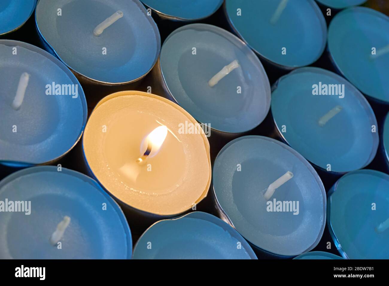 Closeup of tea lights with one being lit. A single warm orange flame between candles in blue shadow. Christmas, mourning or condolence concept. Stock Photo