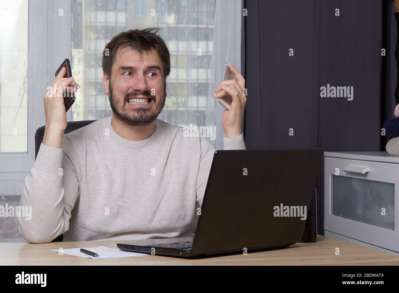 a bearded man is sitting at a table with a laptop in a white sweater looking for a job calling on a mobile phone Stock Photo