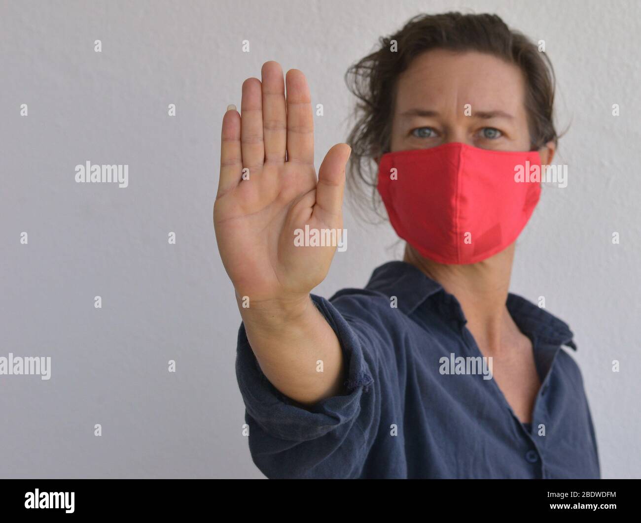 Caucasian woman wears a home-made DIY face mask and makes the hand palm stop sign during the 2019-20 corona-virus pandemic - stay away. Stock Photo