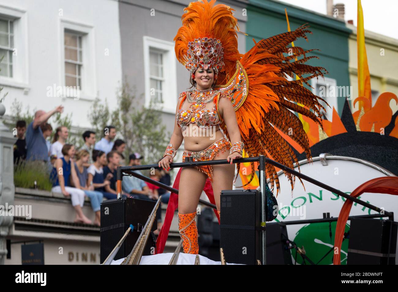 Female performer in orange costume standing atop a float at Notting