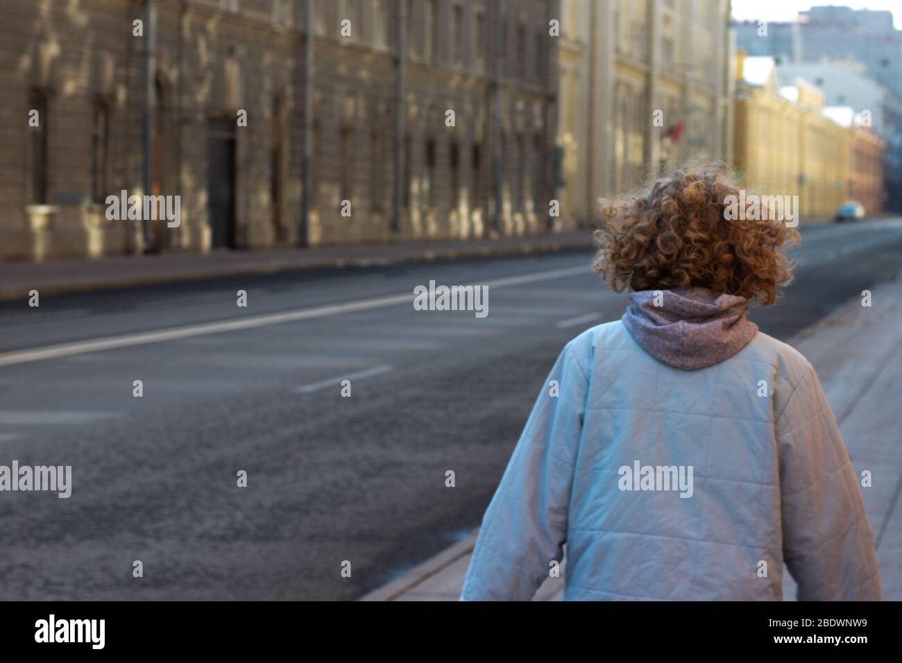 Girl with curly blond hair walks in city street near the road. Copy space outdoor. Photo from the back Stock Photo
