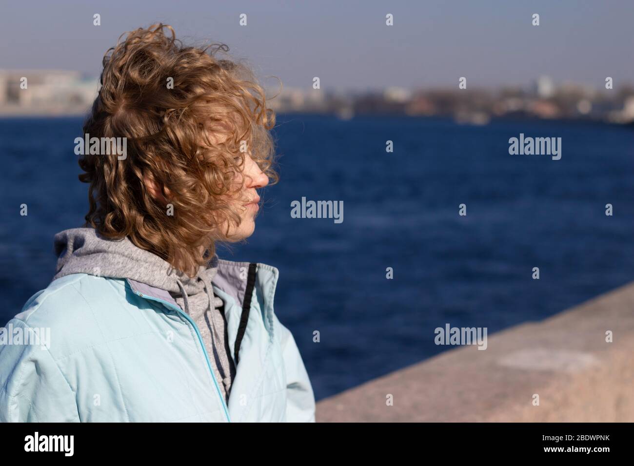 Side view of young girl with blonde curly hair waving in the wind. Copy space on water background Stock Photo
