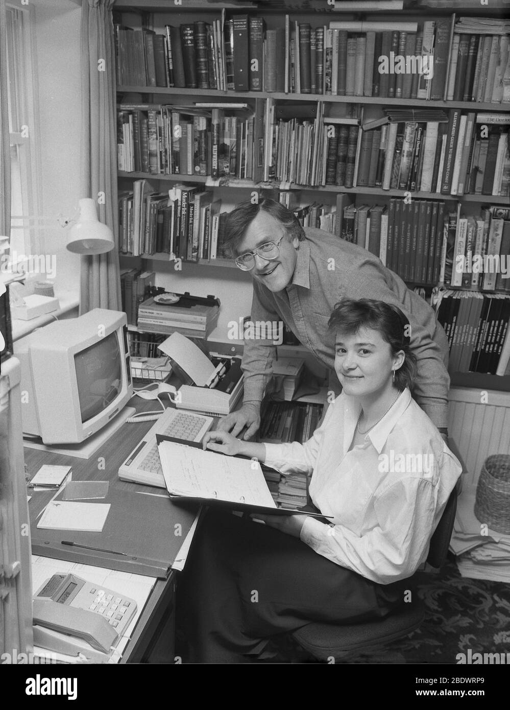 1987, historical, a couple working in a home office. In a compact book-lined study, technology seen on the desk... a computer of the day, keyboard and printer, and telephone. The introduction of the personal computer or PC at this time meant that working from home was far more possible than before. The year 1987 saw the release of the microsoft Windows 2 operating system which introduced a number of popular computing concepts, including windows, desktop icons and colour graphics. Stock Photo