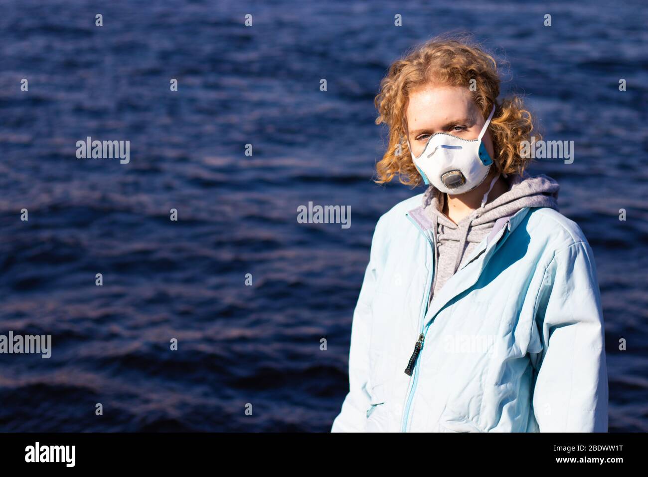 Girl in medical face mask for breath protection looking in camera. Female teen wears respirator. Copy space on background with water Stock Photo