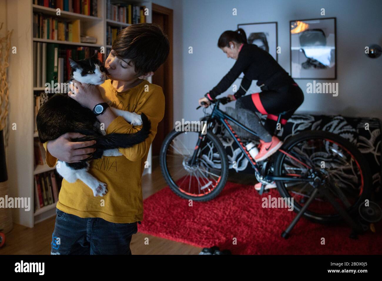Woman doing exercise at home on bike trainer with son in foreground holding their cat during Covid19 confinement in Girona, Catalonia, Spain Stock Photo