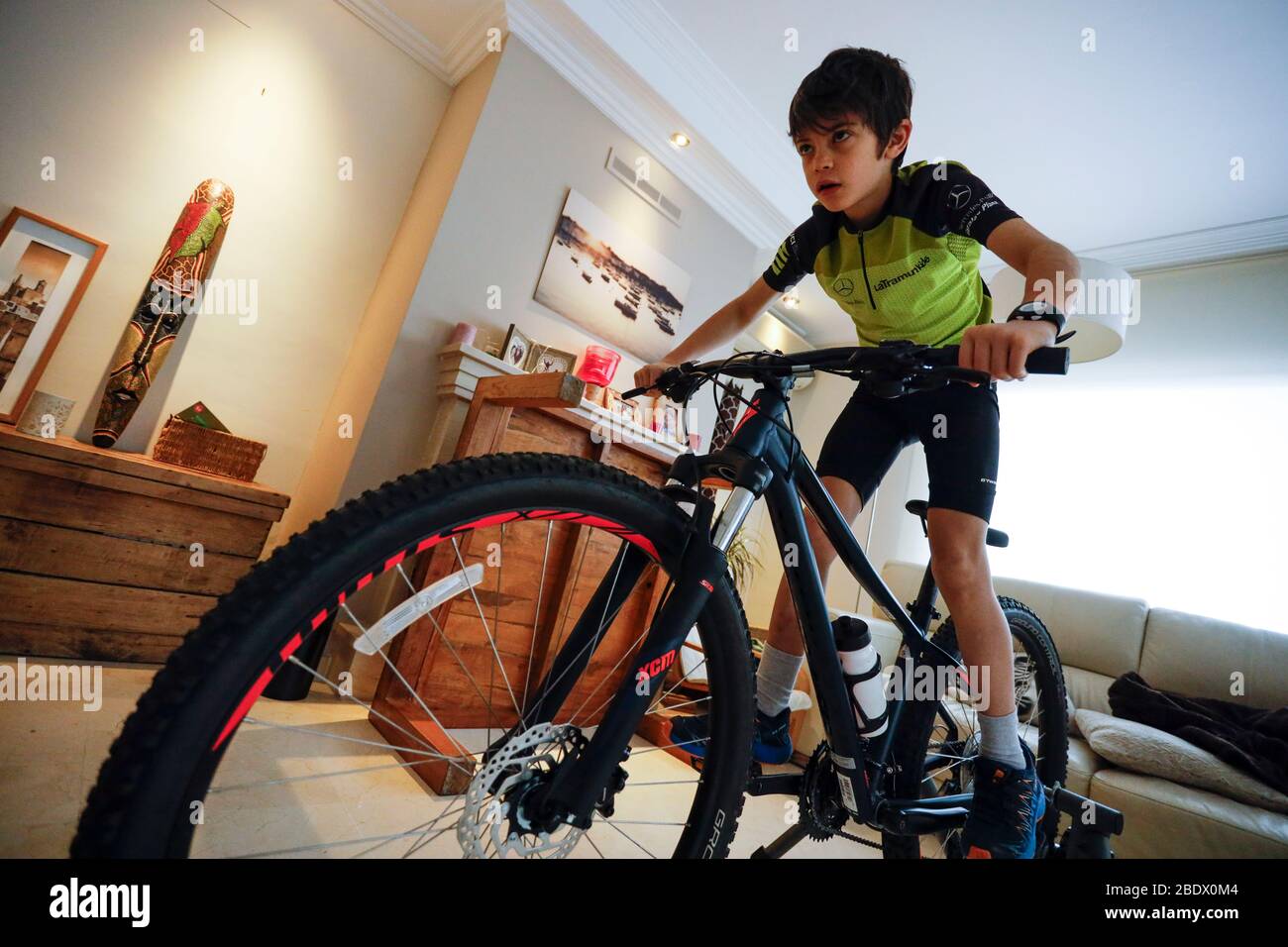 Boy riding bike on a trainer whilst watching television in his house during Covid19 confinement in Girona, Catalonia, Spain Stock Photo