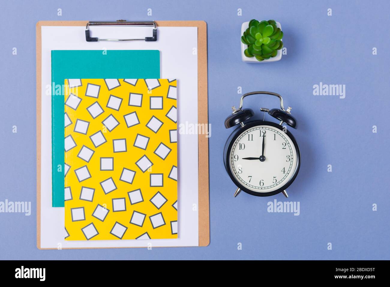 Pile of bright notebooks, clipboard, alarm- clock, table plant and stationary on blue desk top. Copy space. Free space. Start of working day.  Stock Photo