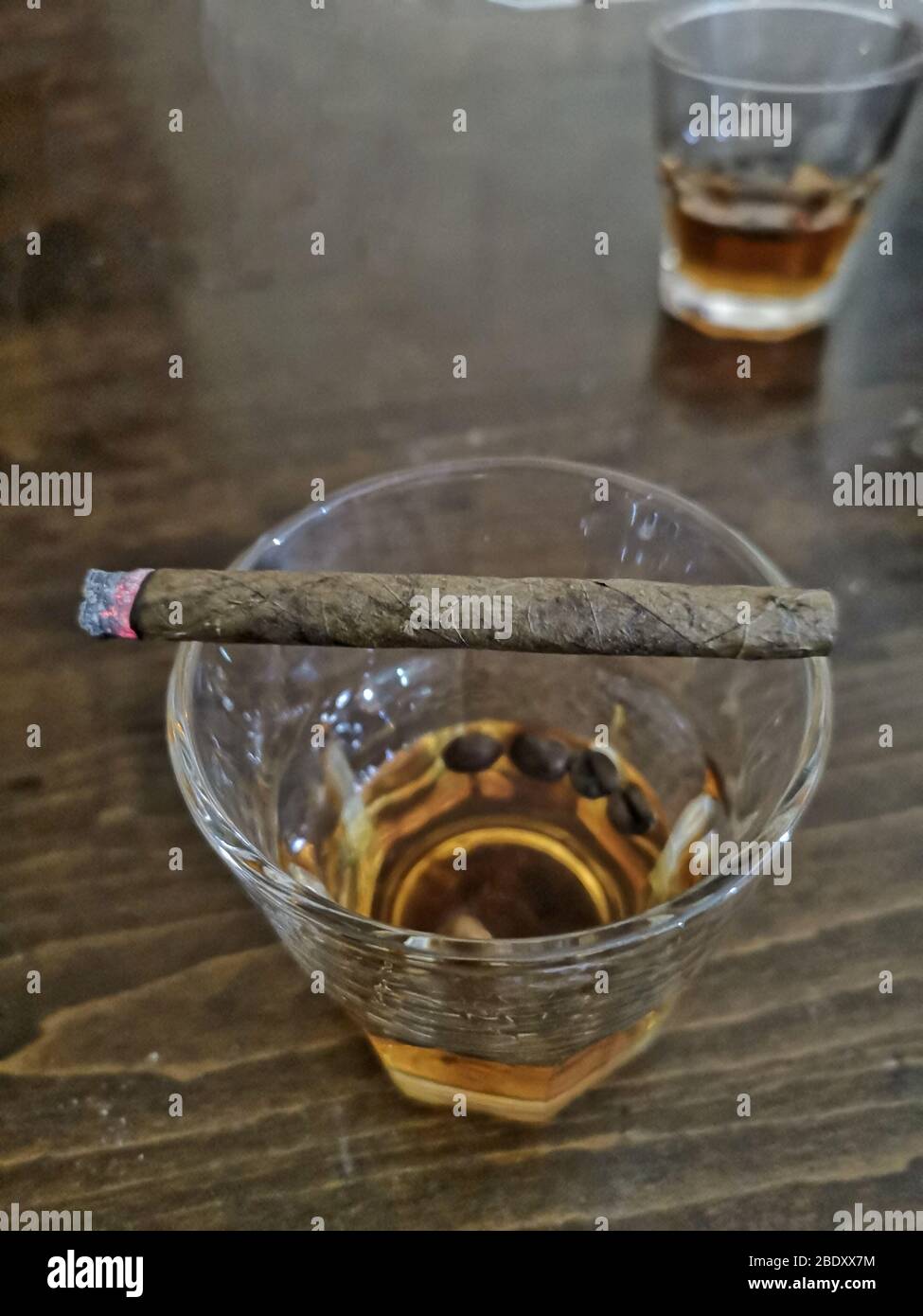 Close up shot of half full glass of whisky on top of wooden bar with lit cigar on top of it, why smoking and alcoholism is bad, sharing scotch whiskey Stock Photo