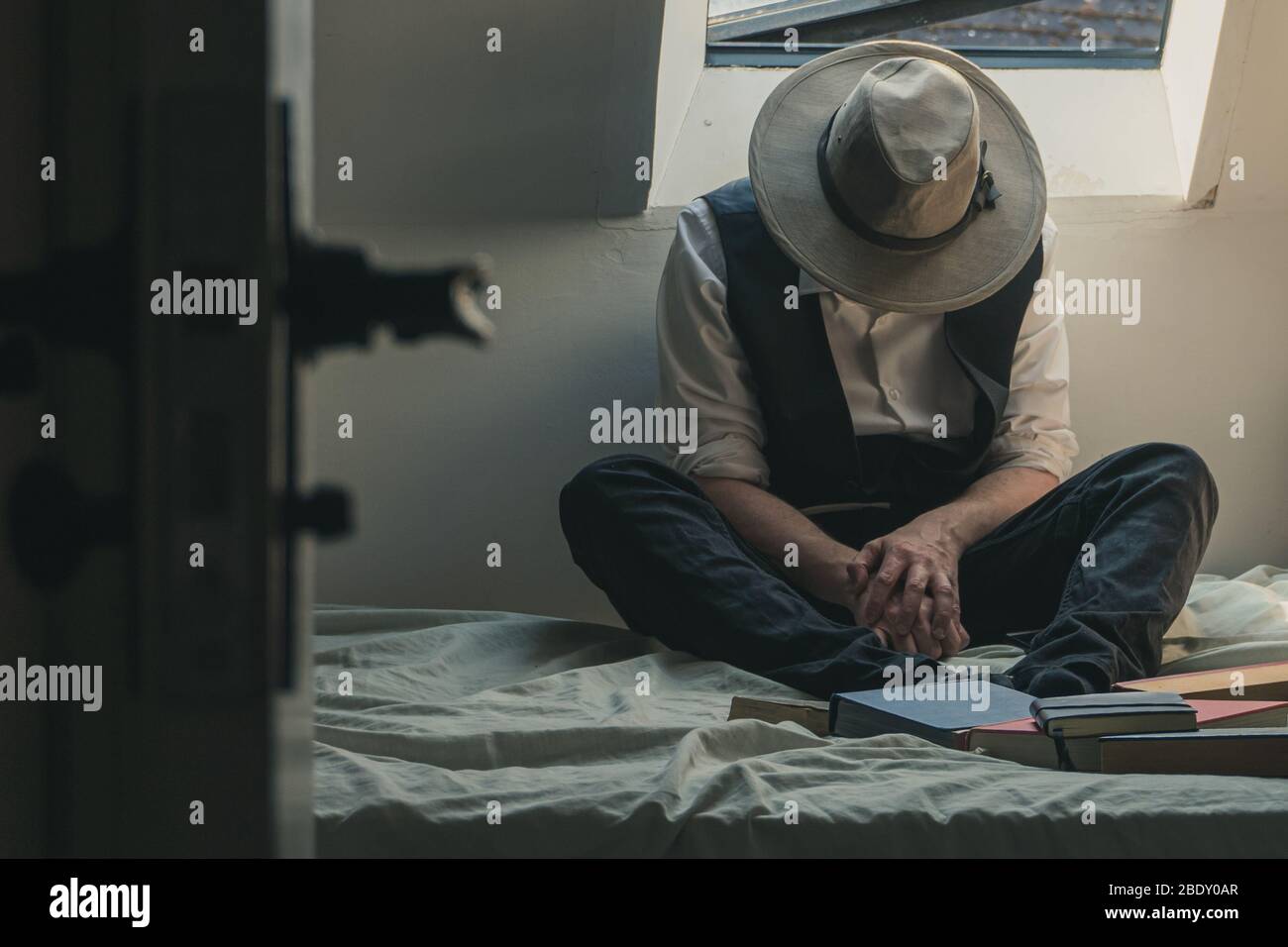 A man looking down wearing a fedora hat and waistcoat sitting on a bed with books surrounding him. Stock Photo