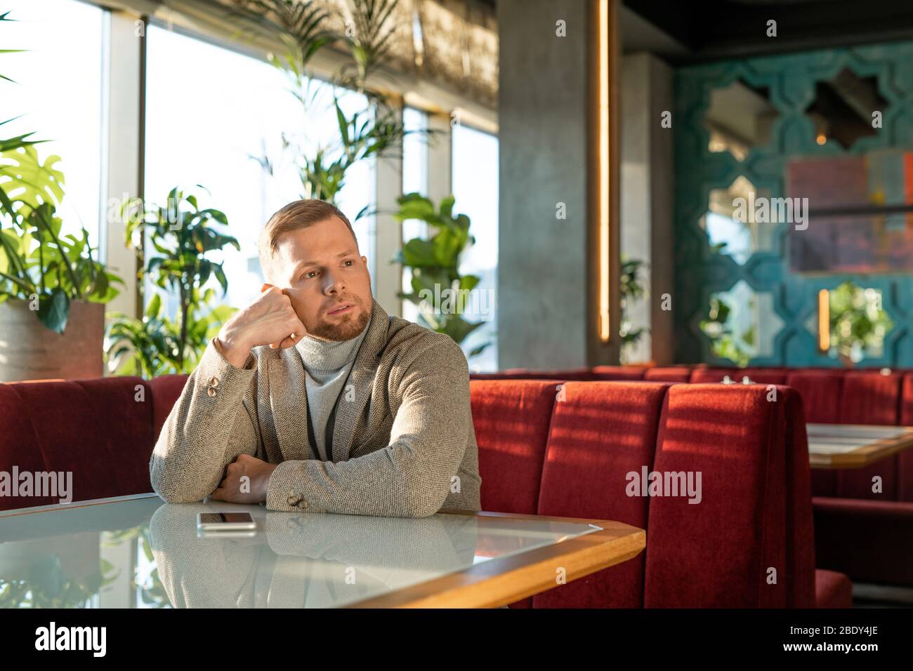 Young pensive man in jacket and pullover sitting on red velvet couch by table in classy restaurant and waiting for his girlfriend Stock Photo