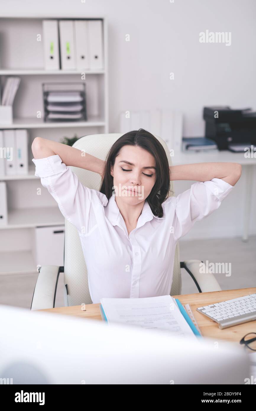 Young serene female office manager relaxing in armchair by desk while keeping hands behind head