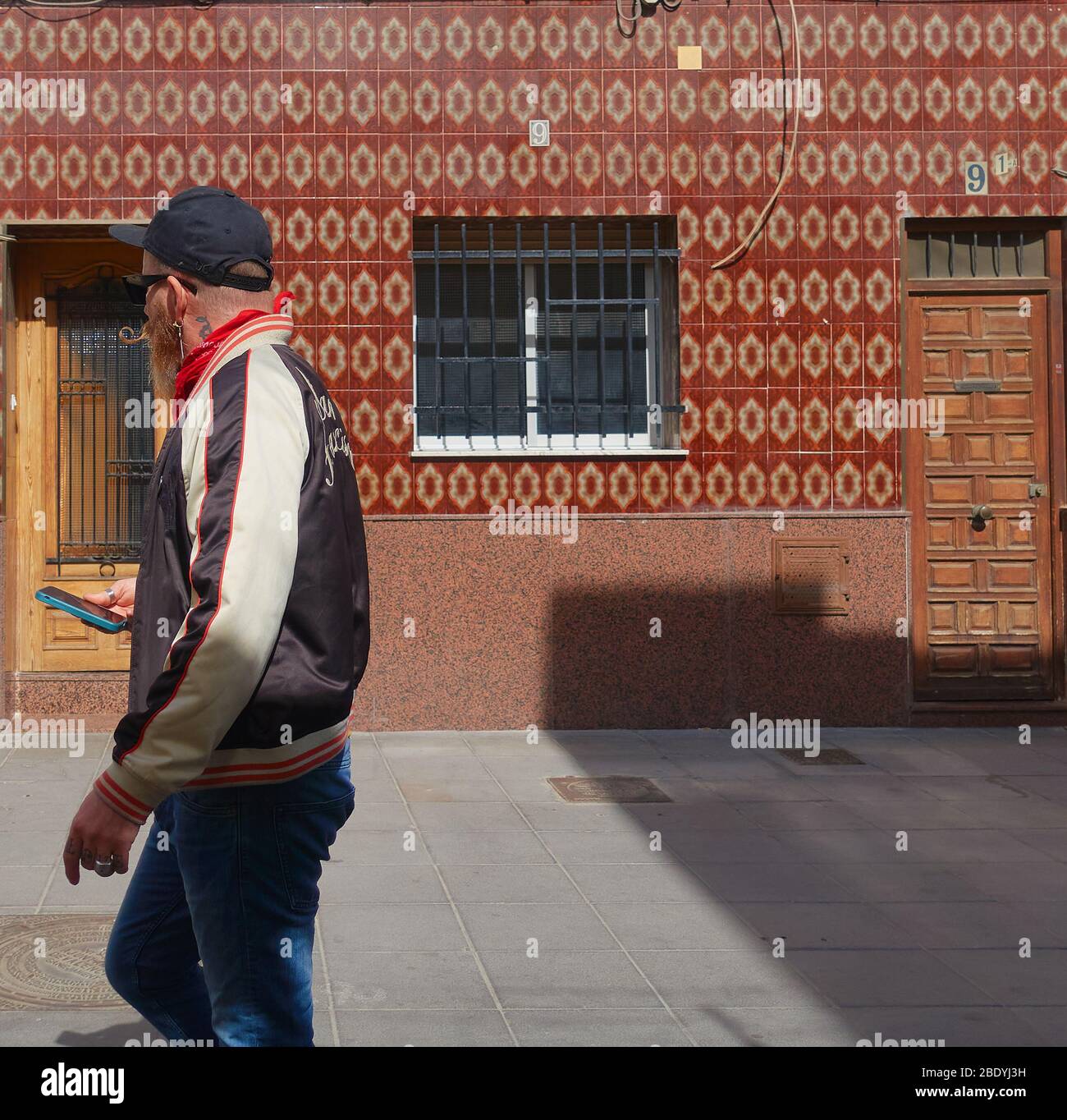 An edgy hipster man walking with his mobile looking away from camera as he passes a traditional tiled house in El Cabanyal, Valencia, Valencia, Spain Stock Photo