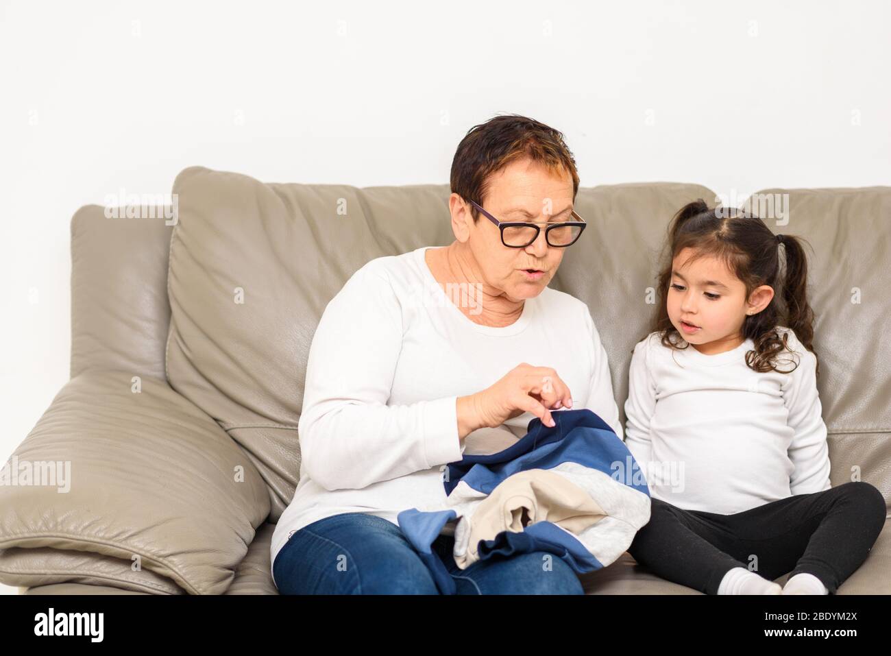 Grandmother Teaches Her Granddaughter To Mend Clothes. Kid Being Homeschooled Due To School Shut Down During Covid-19 Pandemic. Stock Photo