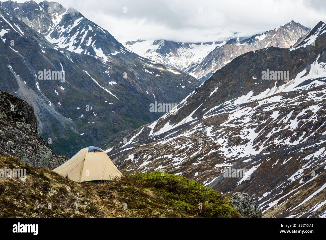 Tan and grey tent on narrow ledge of tundra and rock overlooking a large valley. Deep in the wilderness of the Talkeetna Mountains of remote Alaska. Stock Photo