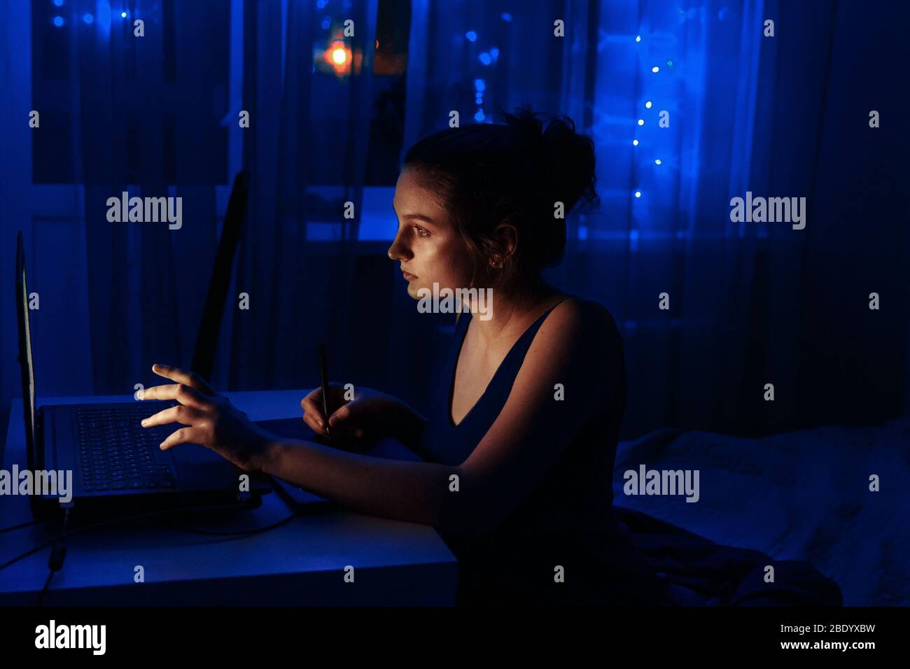 A young girl works remotely from home. The girl is sitting at the computer and drawing on a tablet. Stock Photo