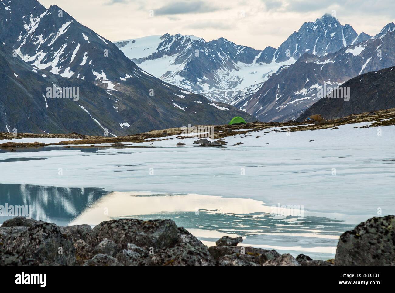 Small green one-person tent above mostly frozen lake in the Alaskan wilderness. Beyond camp are snowy glaciers and craggy summits. Stock Photo