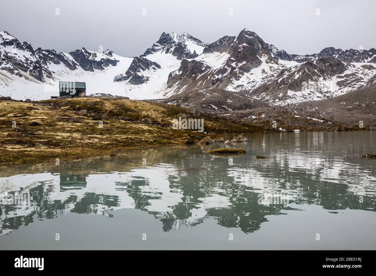Small green backcountry hut in remote Alaska, reflected in a calm alpine lake in the Talkeetna Mountains. Stock Photo