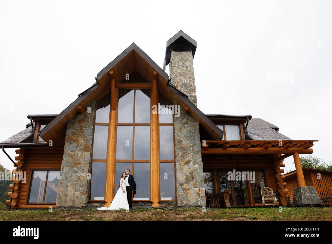 A woman in a wedding dress and a man in a stylish suit stand in front of a gorgeous house in the mountains Stock Photo