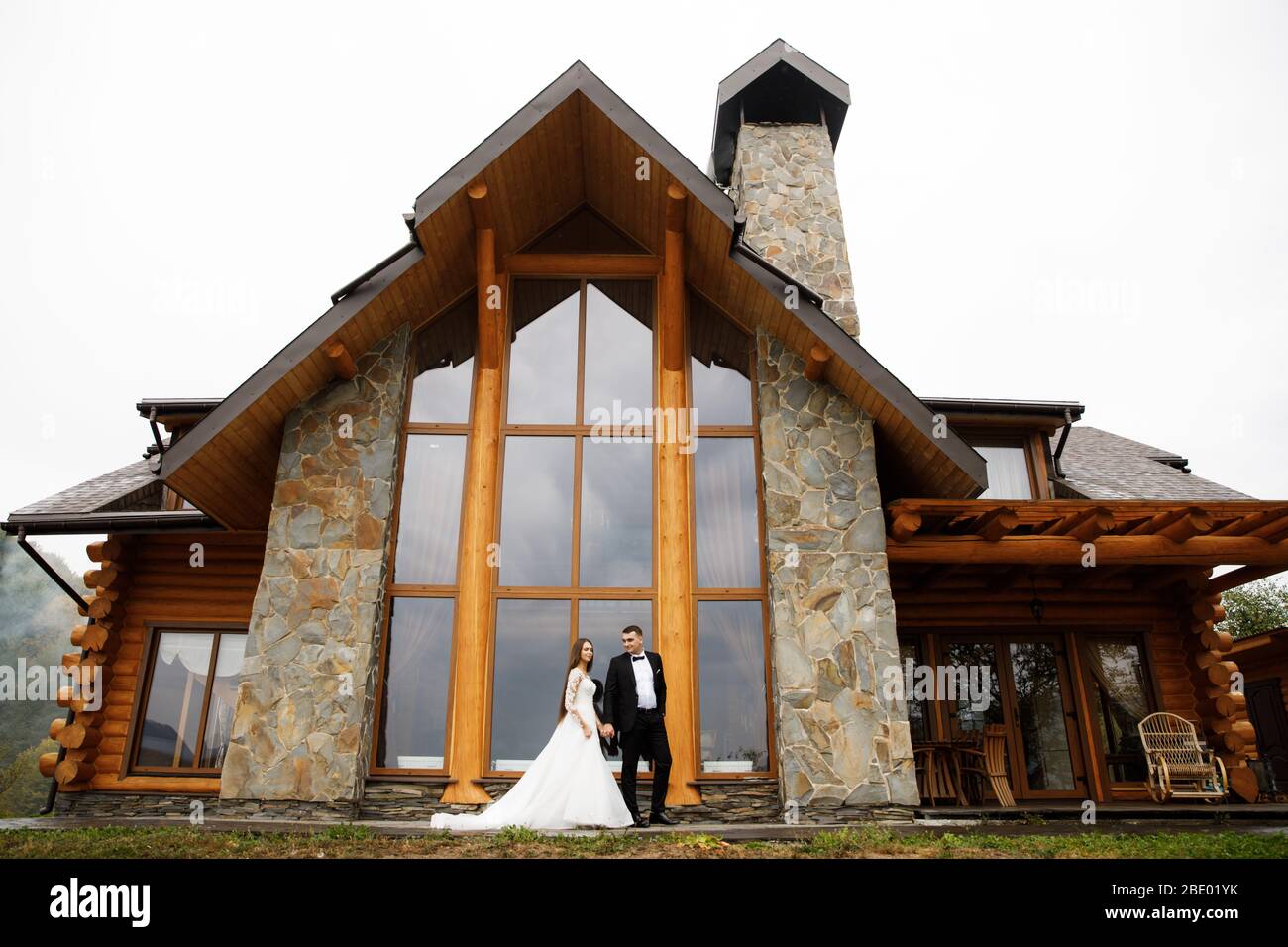 A woman in a wedding dress and a man in a stylish suit stand in front of a gorgeous house in the mountains Stock Photo