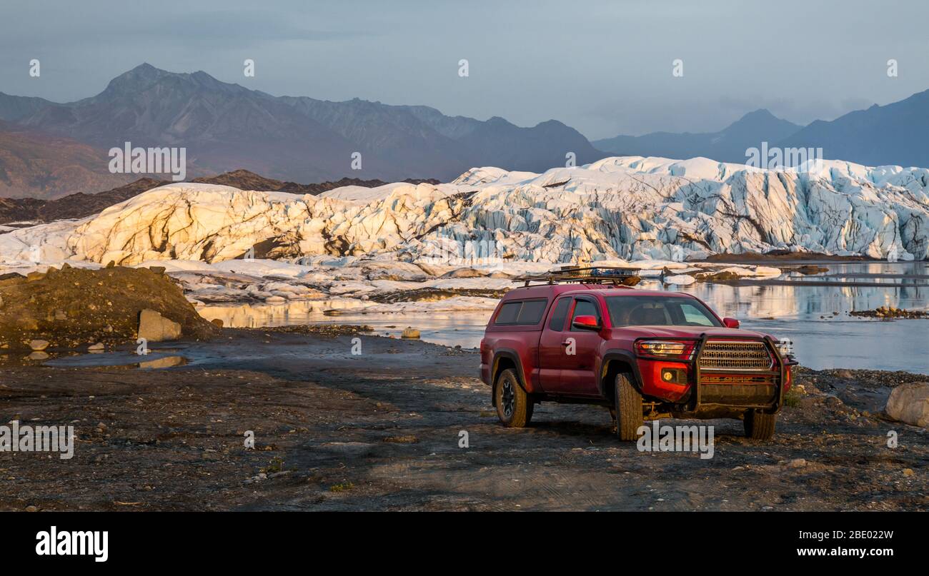 Red pickup truck camper parked in front of a glacial lake and icefall. Offroad driving in remote Alaska. Stock Photo