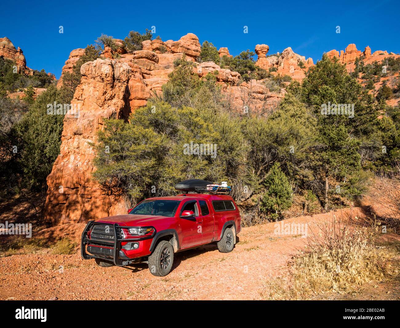 Orange sandstone towers stand above a narrow dirt trail where a red pickup camper is parked. The truck is loaded with camping and overland gear for of Stock Photo