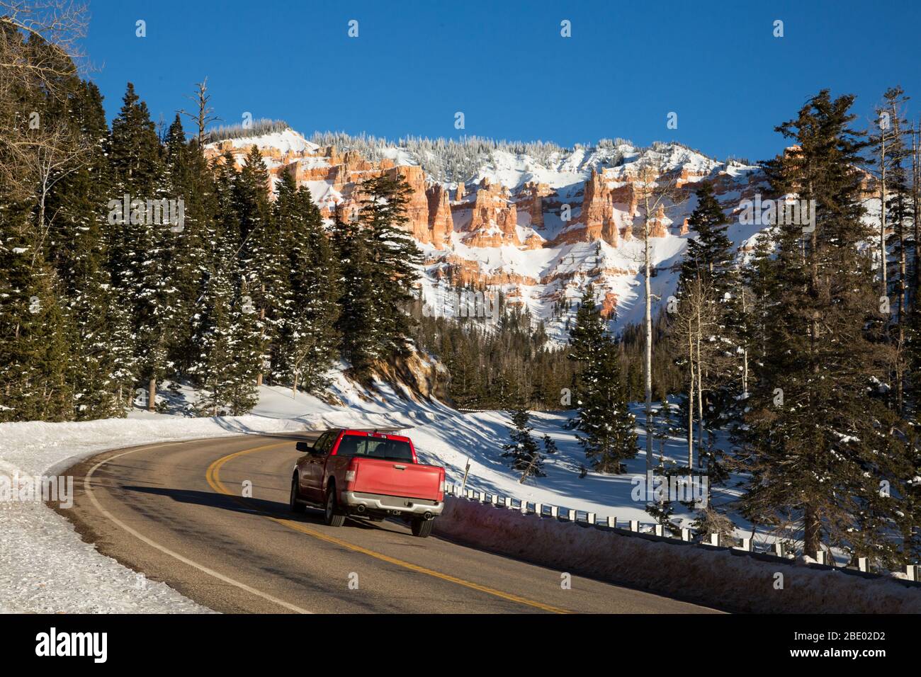 Red pickup driving up a winding canyon road toward snow covered rock formations in the red and orange sandstone. Stock Photo