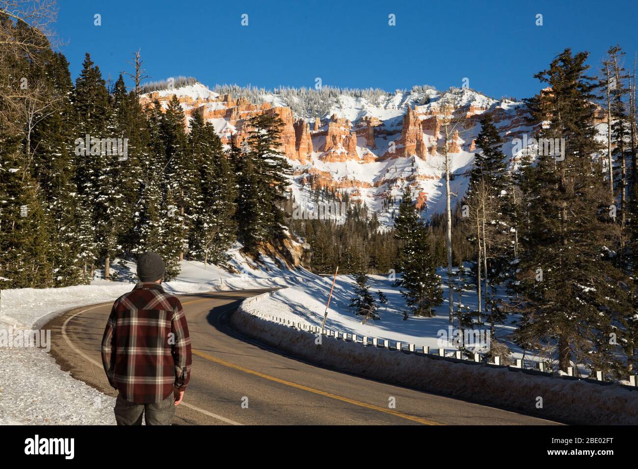 Man in red plaid shirt and warm cap standing on the side of a highway. The road leads toward a sandstone rock formation of red and orange towers cover Stock Photo