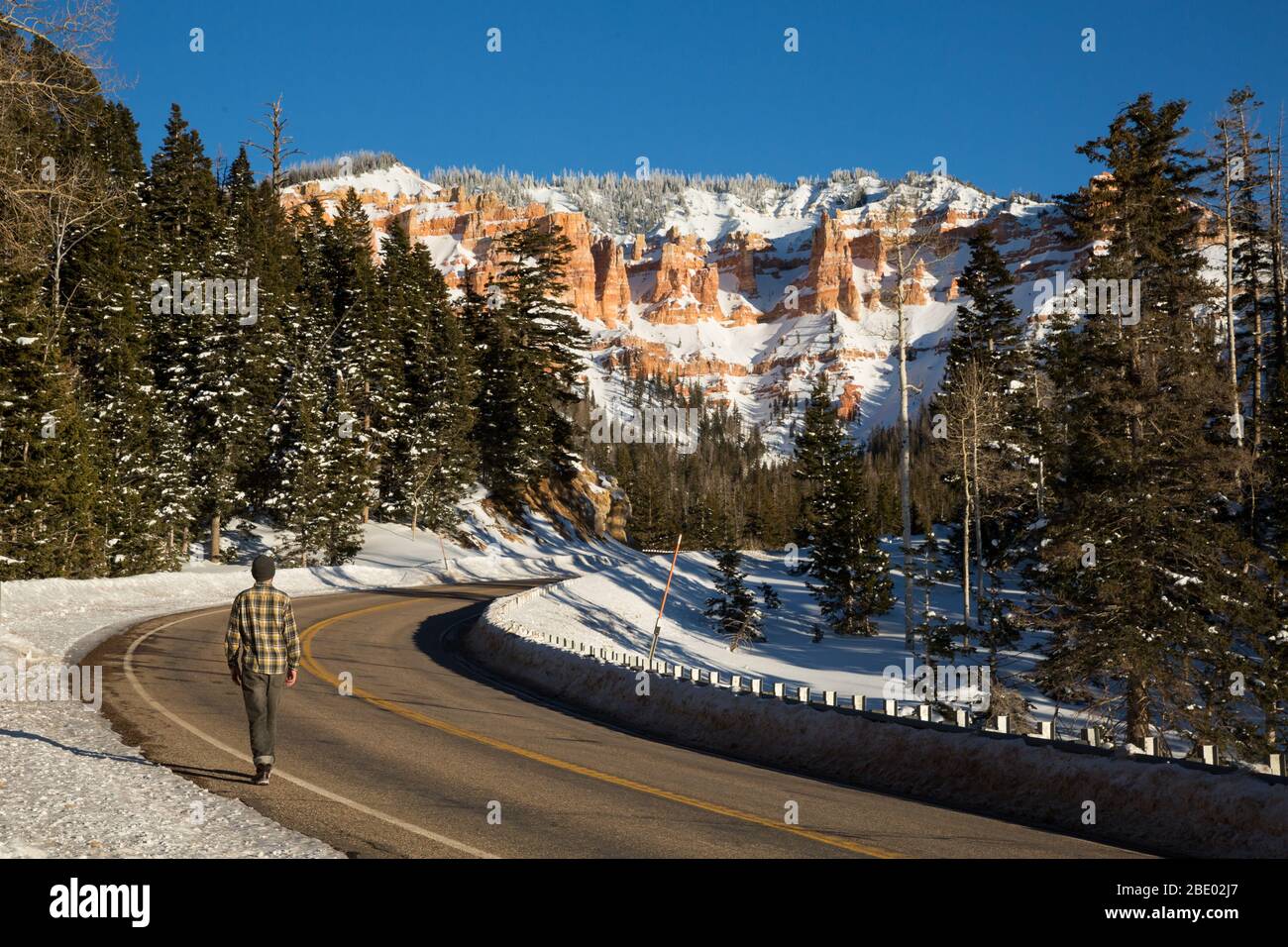 Man walking beside a two-lane road in the winter. Overhead are desert rock formations covered in snow. Stock Photo