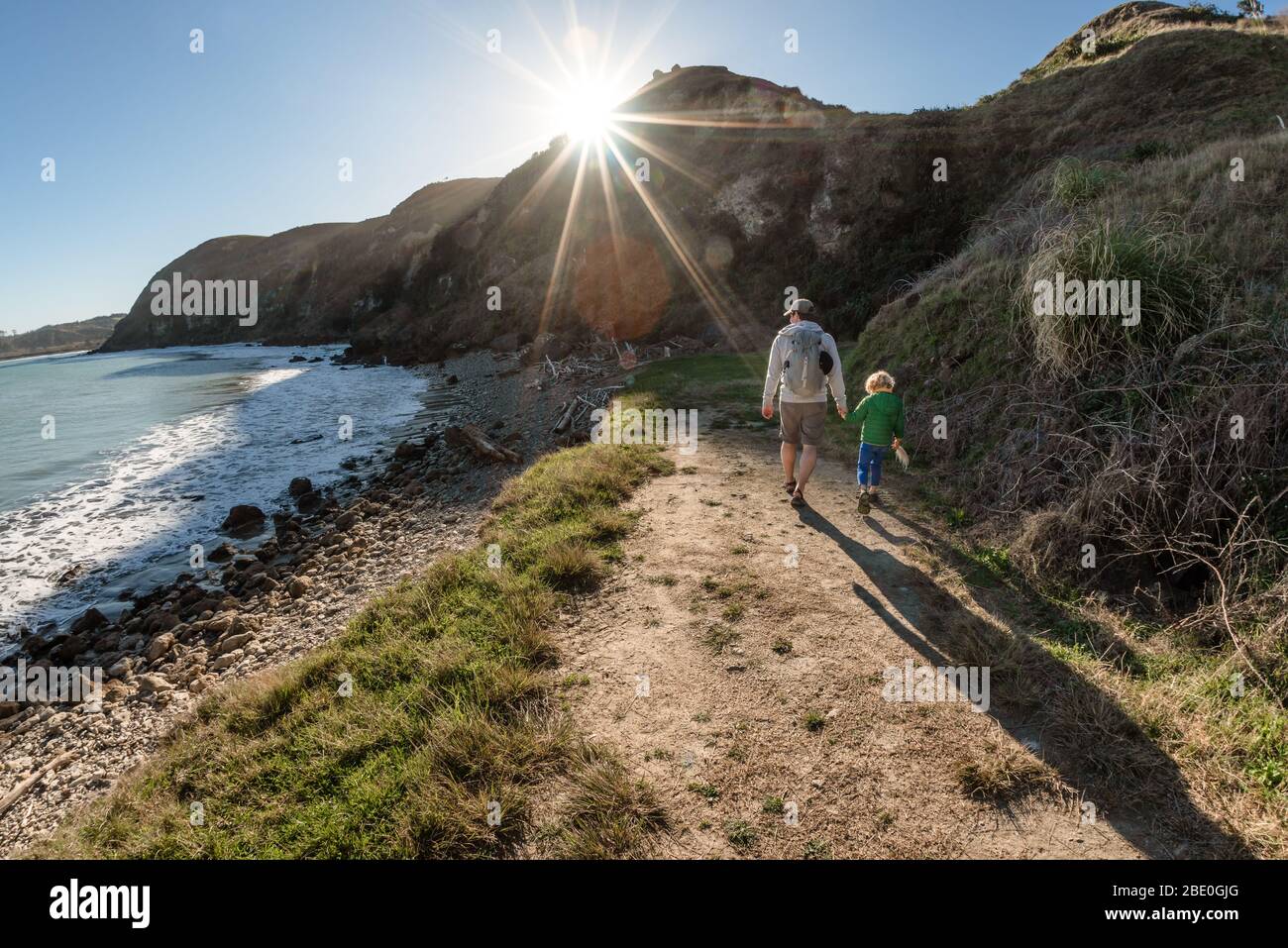 Dad and small child holding hands on path near ocean on a sunny day Stock Photo