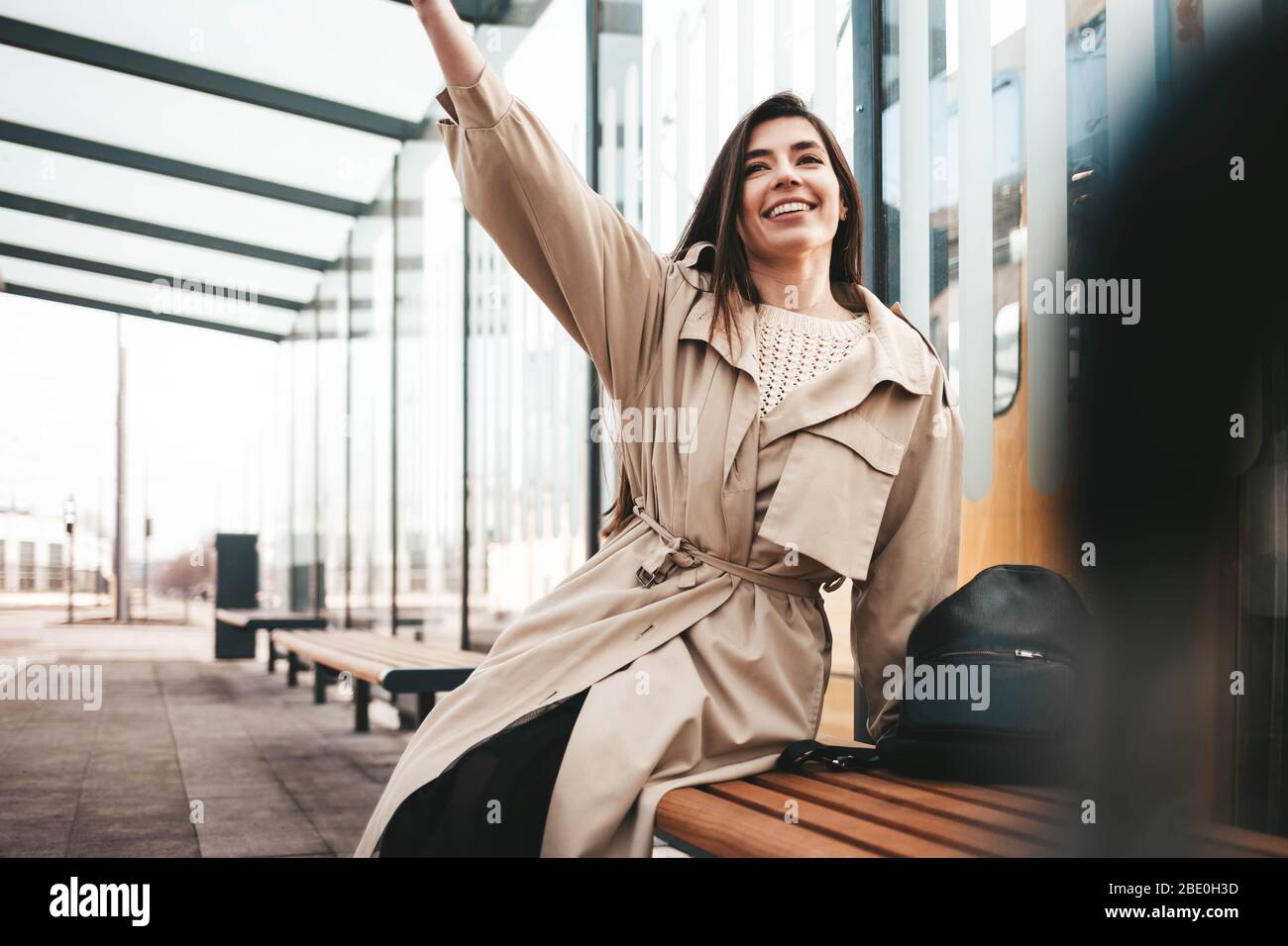 Positive young woman sitting at a public transport stop and waving Stock Photo