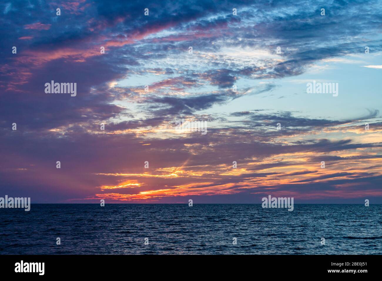 Pink sky and light rays shining through clouds as sun sets on ocean. Stock Photo