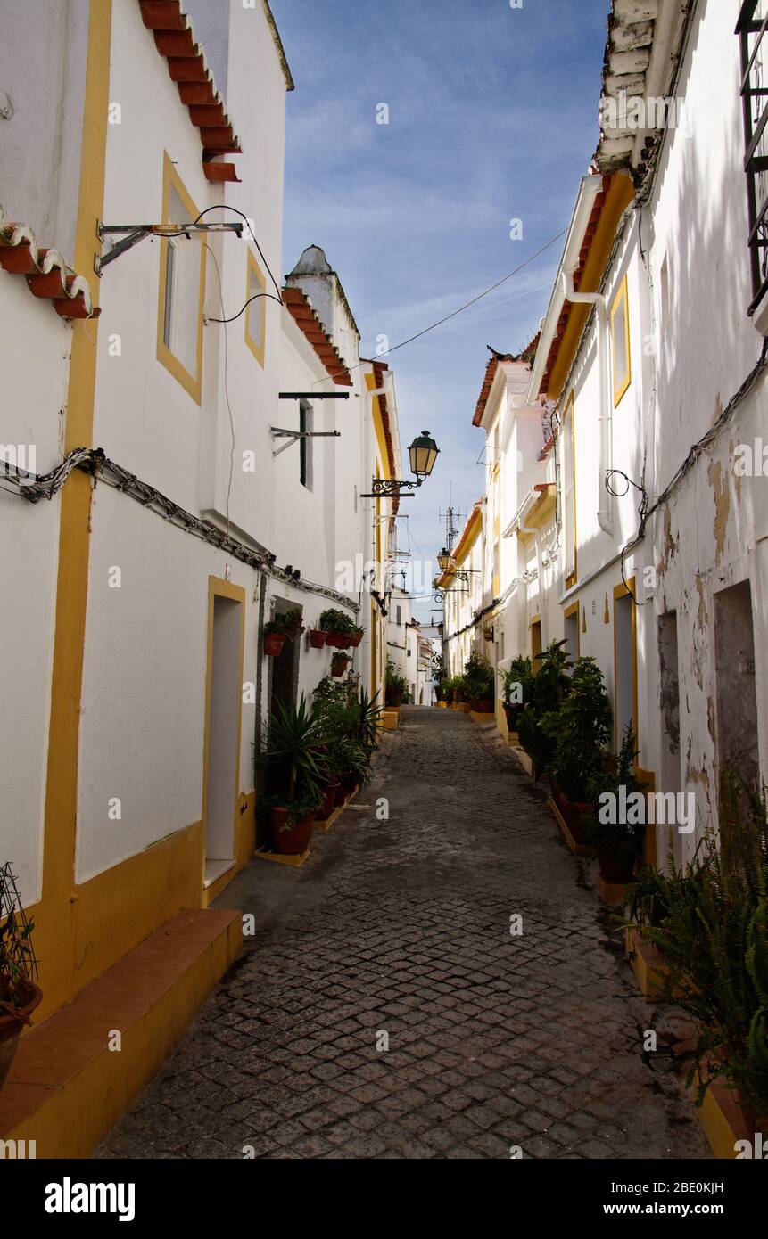 Narrow typical cobblestone street of white walls and yellow bordering of the jewish borough at Elvas. Light lamps and blue sky. Portugal. Stock Photo