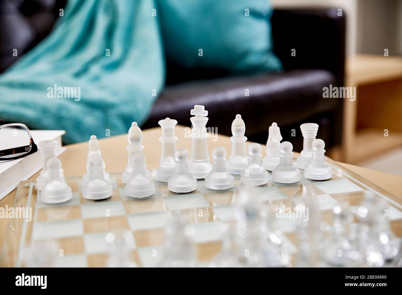 Close up of frosted glass chess pieces on a glass chess board in a living room with reading glasses and a hardcover book and shallow depth of field Stock Photo