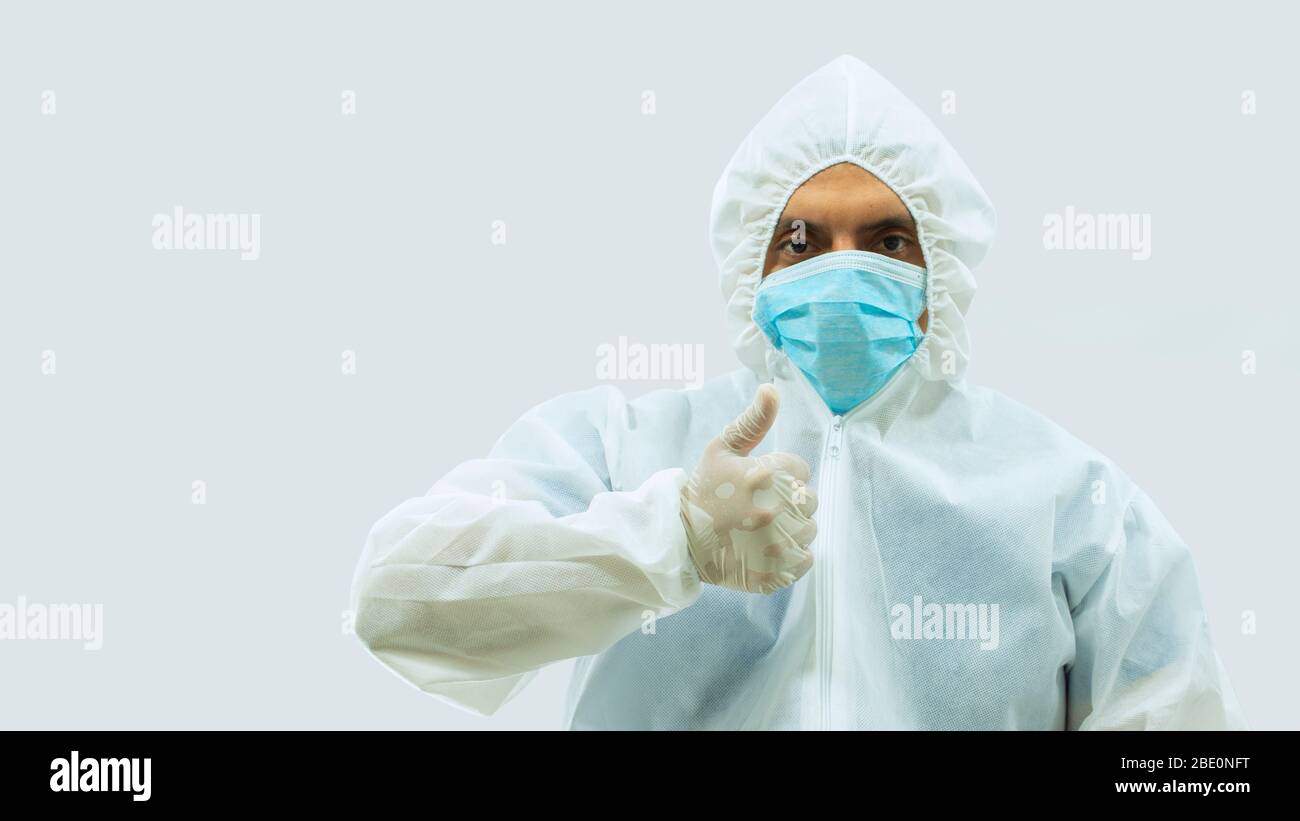 Doctor in white bioprotective suit and blue mask standing with his arm outstretched and his hand with thumb up making LIKE sign on white background Stock Photo