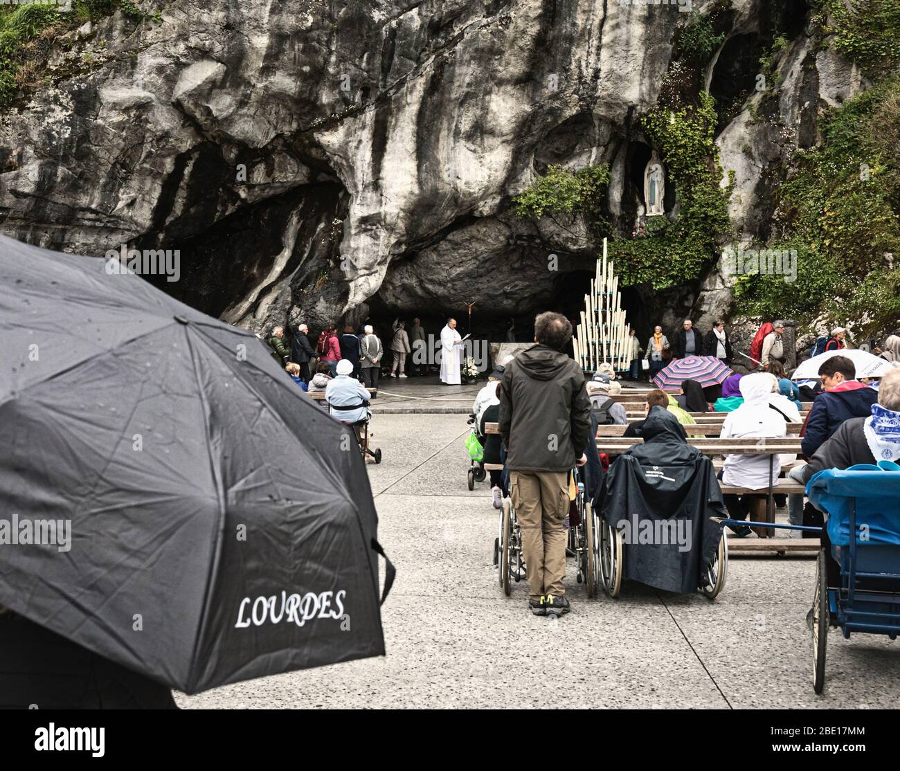 A priest recites prayers at the entrance to the Massabielle grotto of the Sanctuary of Our Lady of Lourdes, while the faithful queue to enter.  It was Stock Photo