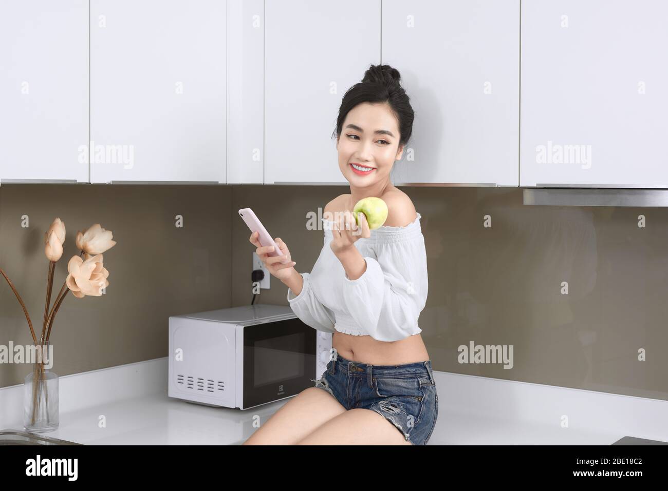 Smiling pretty Asian woman looking at mobile phone and holding an apple in kitchen. Stock Photo