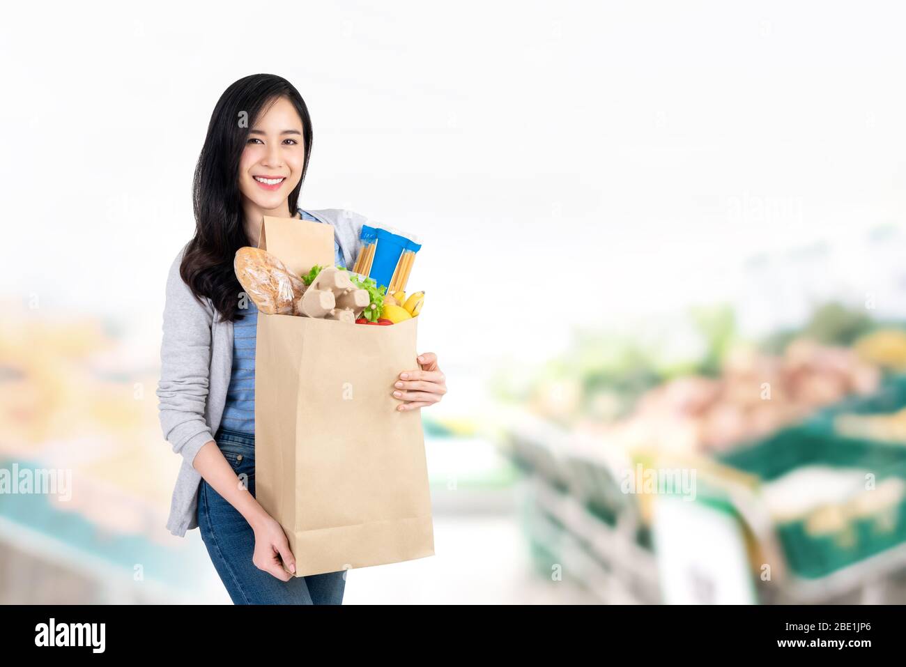 Beautiful smiling Asian woman holding paper shopping bag full of groceries in supermarket background with copy space Stock Photo