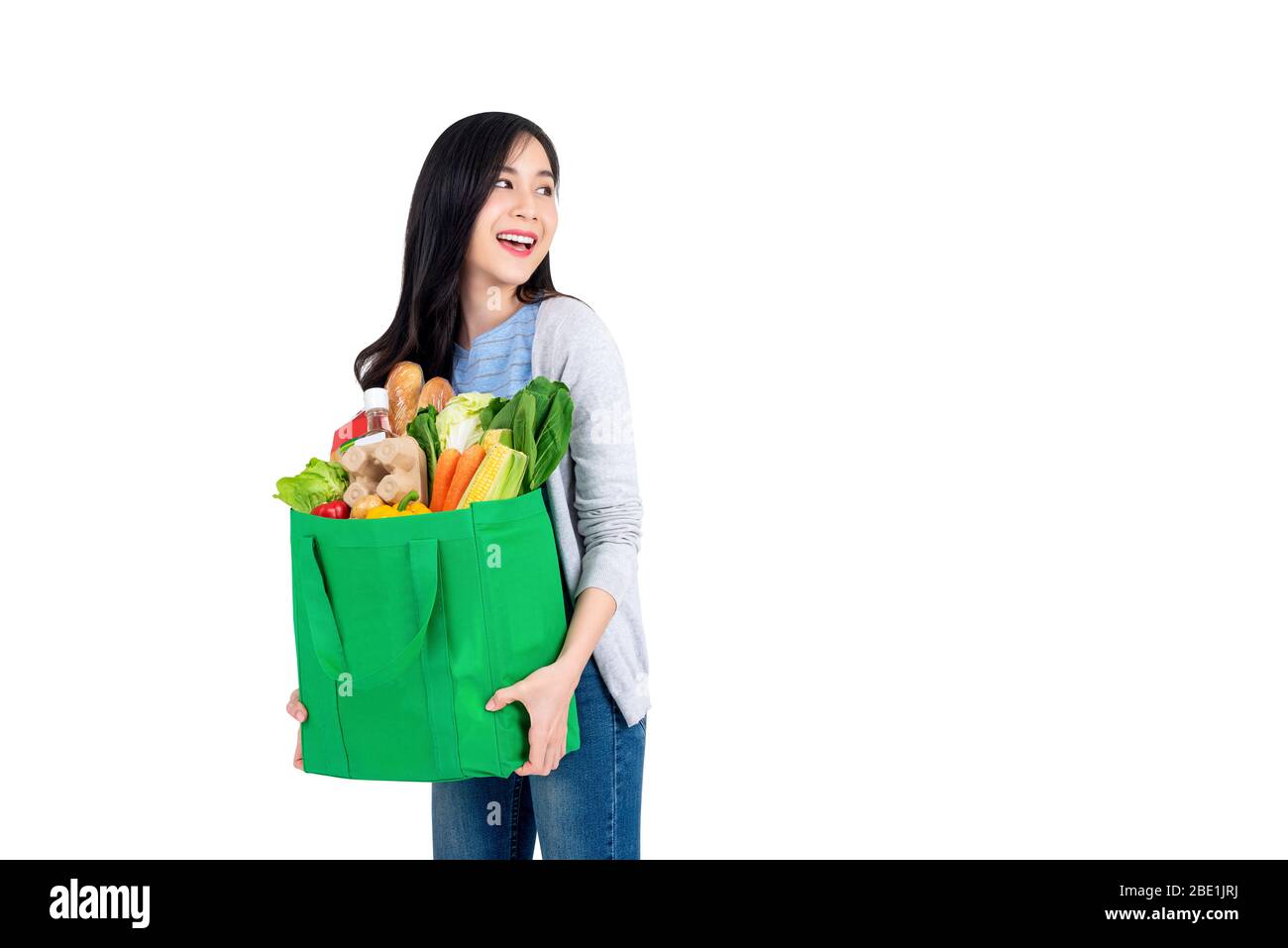 Beautiful smiling Asian woman holding reusable green shopping bag full of groceries and looking to copy space aside isolated on white background Stock Photo