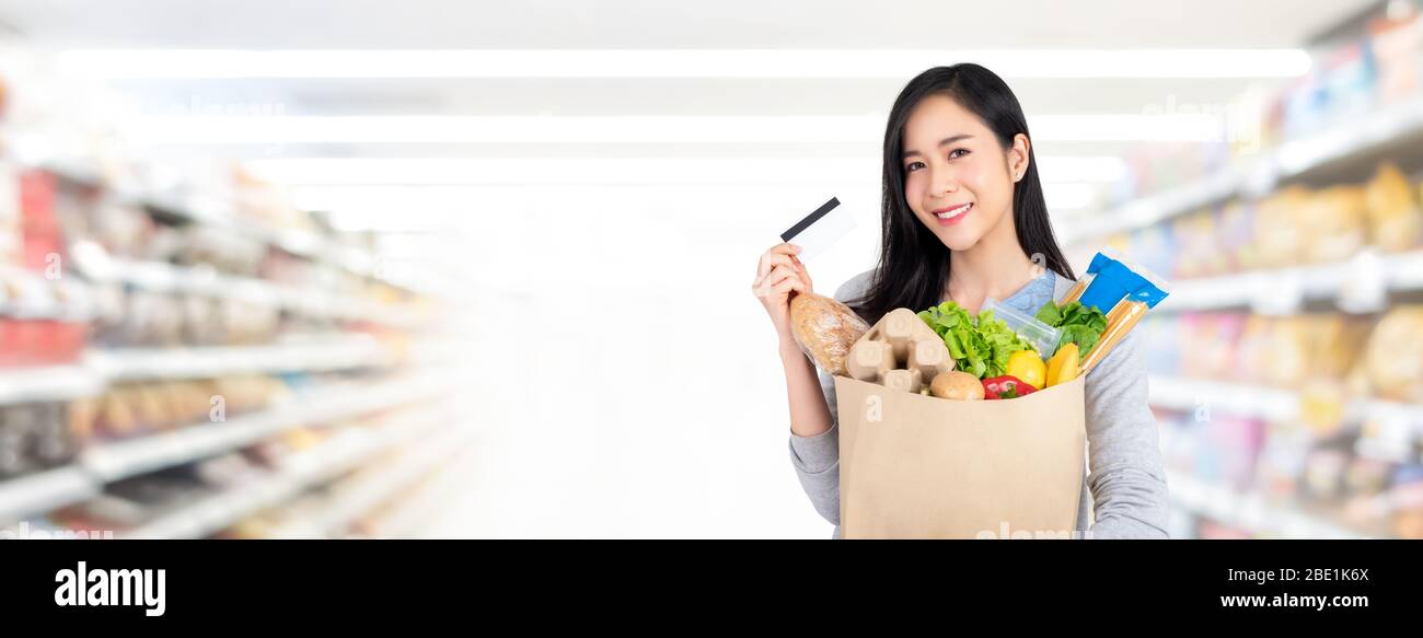 Beautiful Asian woman holding paper bag full of groceries shopping with credit card in supermarket banner background with copy space Stock Photo