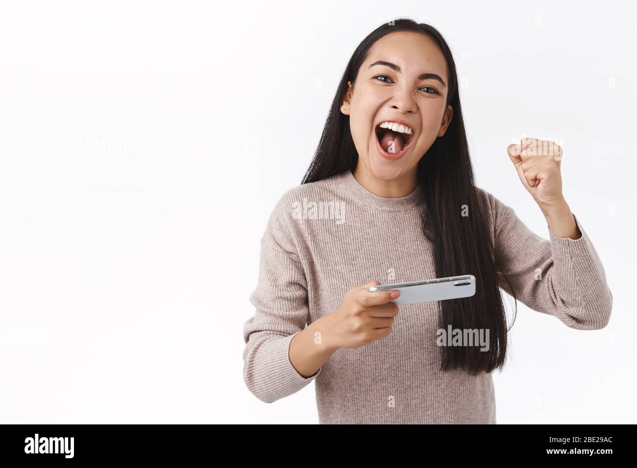 Enthusiastic good-looking cheerful asian young girl winning game, beat the score, fist pump triumphing and yelling yes as encourage herself, holding Stock Photo