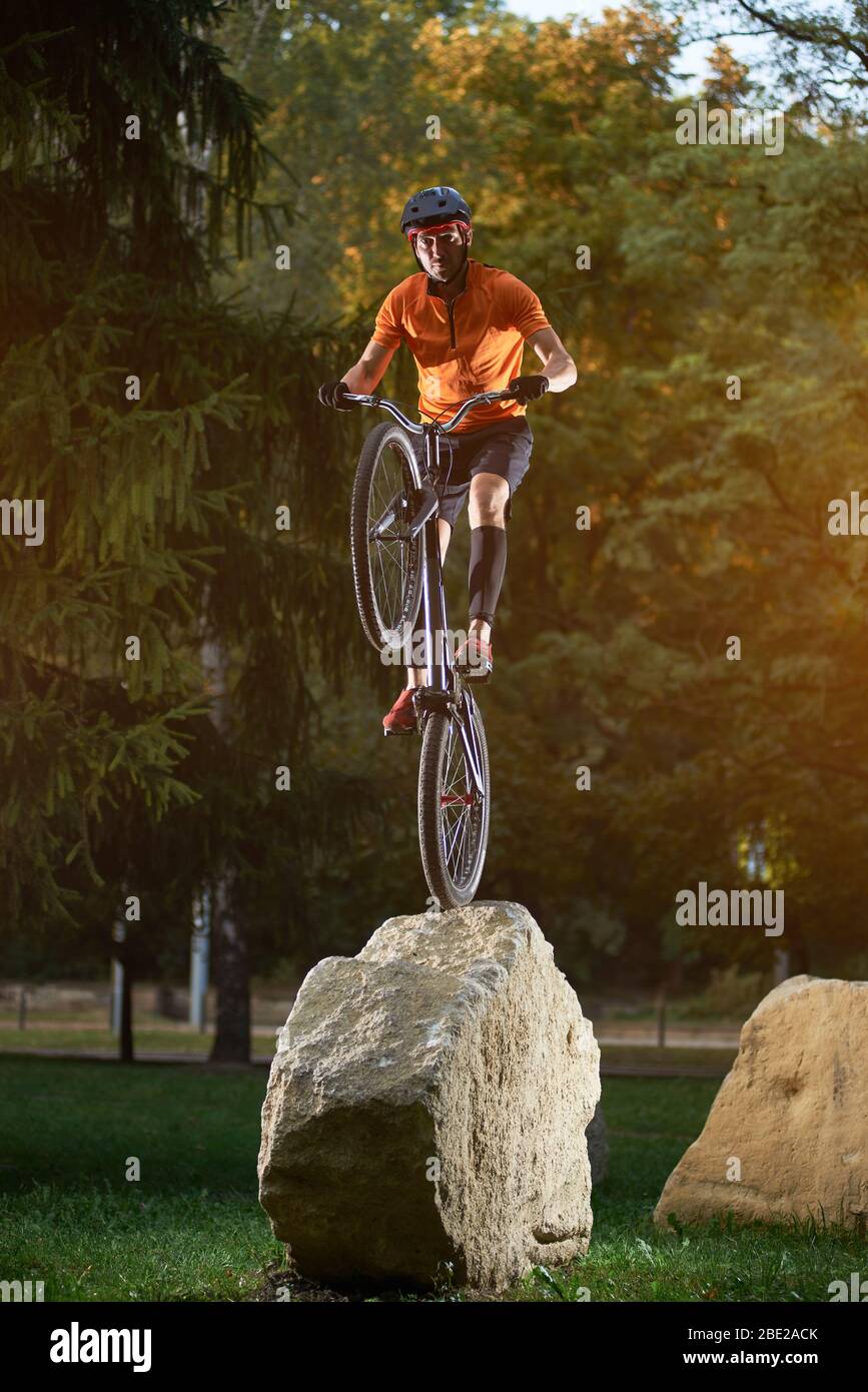 Portrait of young cyclist standing on back wheel of bmx bike on a rock looking at camera in park, green trees on background Stock Photo