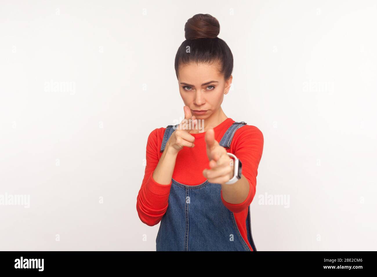 Crime, killing gesture. Portrait of dangerous girl with hair bun in denim overalls imitating guns, shooting into camera with finger pistols, looking m Stock Photo