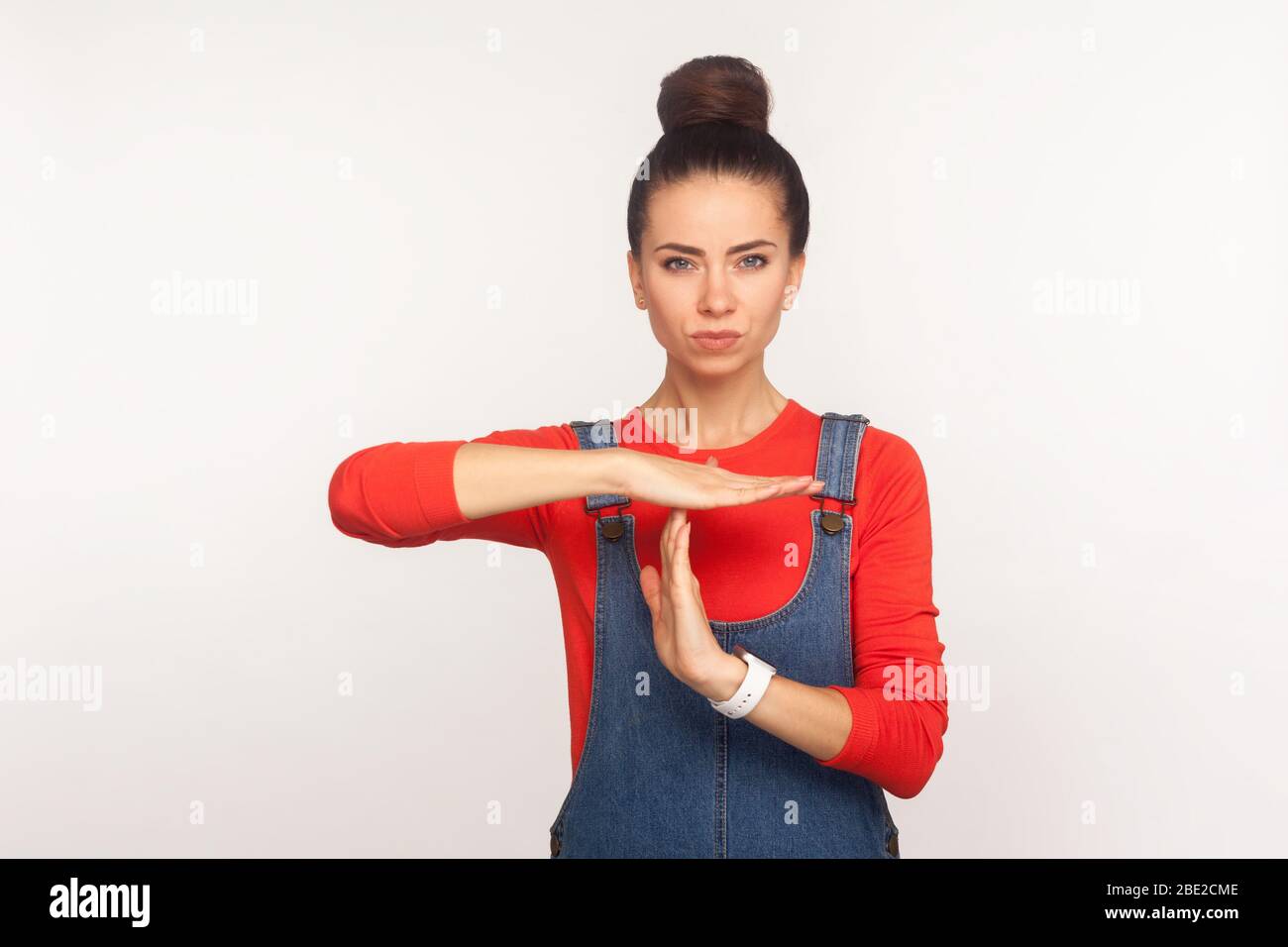 I need break! Portrait of tired displeased girl with hair bun in denim overalls showing time out gesture and looking with dissatisfied expression. ind Stock Photo