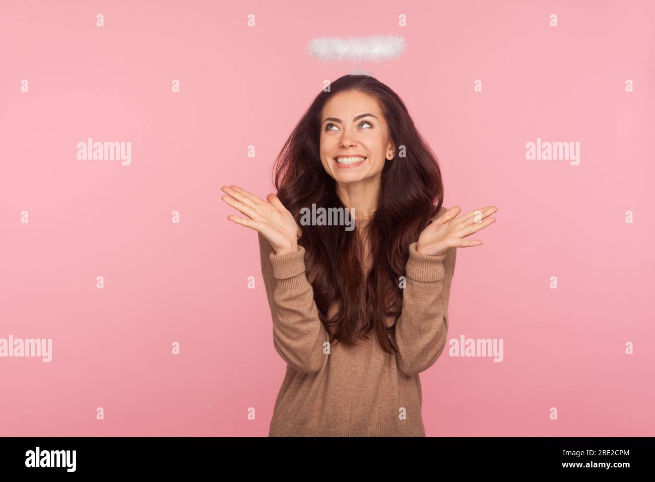 Portrait of lovely playful young woman with halo above head raising hands in bewilderment, gesturing I don't know, looking up and smiling confused. in Stock Photo