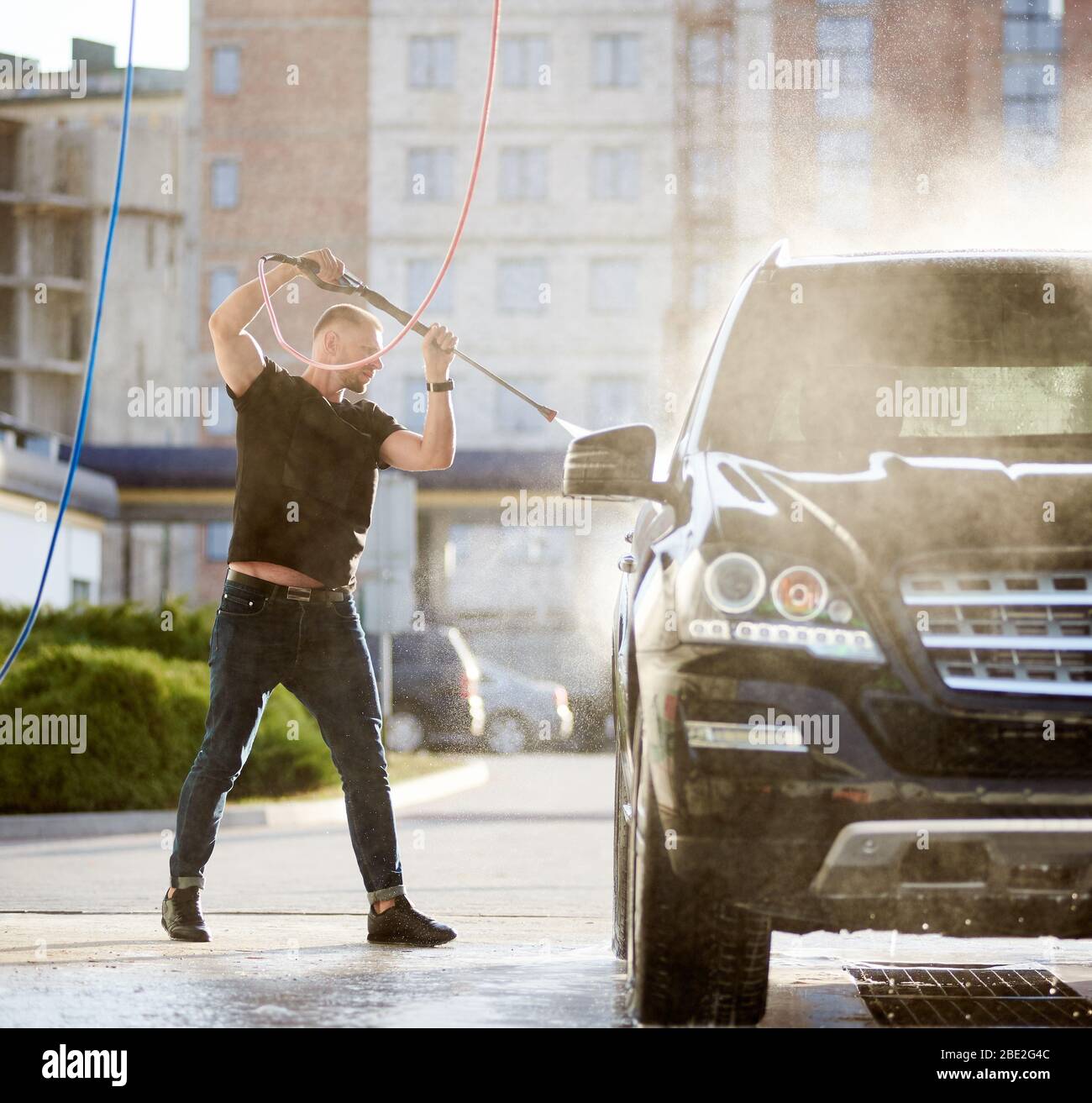 Full length view of a cool man cleaning his black car outdoors, water is splashing over the top of the car shining on the sun, residential building is on the background Stock Photo