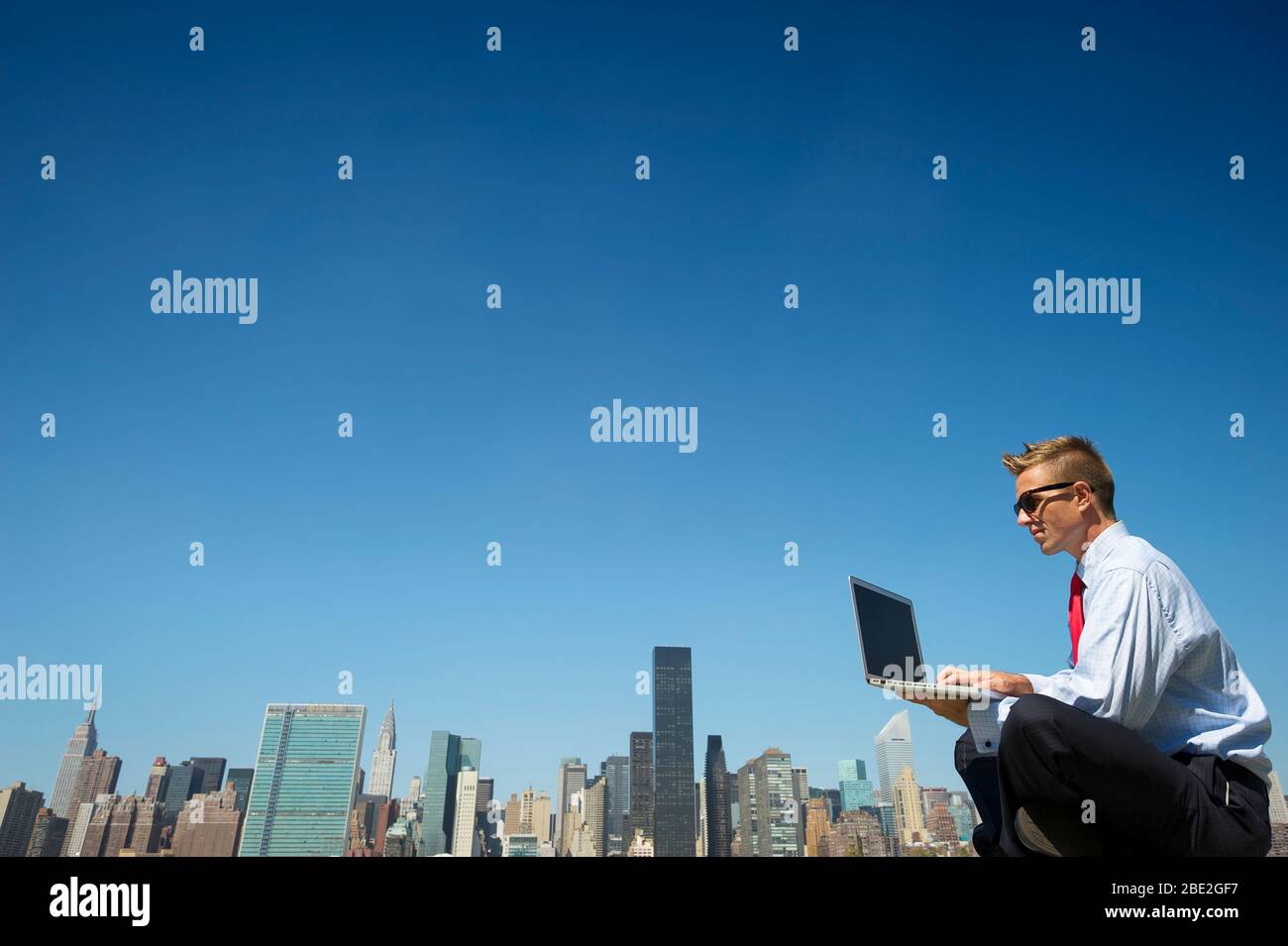 Businessman in a suit sitting outdoors using laptop computer above the city skyline under clear blue sky copy space Stock Photo