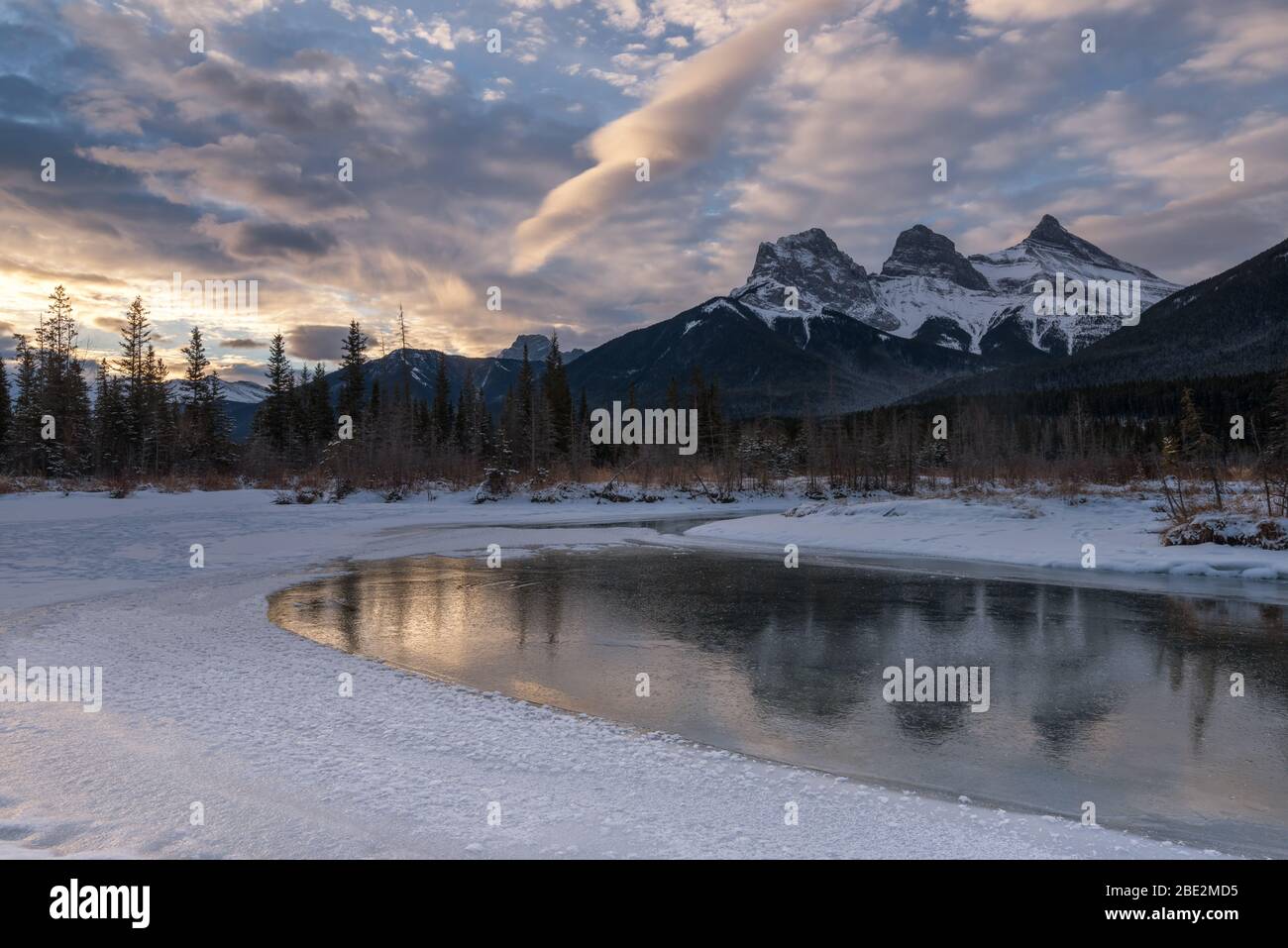 Snow covered Three Sisters peaks reflecting in calm icy water, morning atmosphere, Bow River, Canmore, Banff National Park, Alberta, Canada Stock Photo