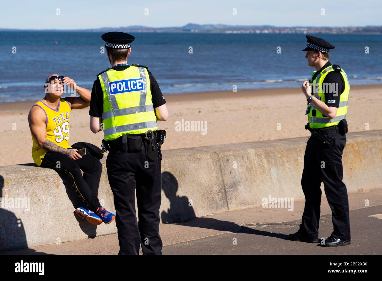 Portobello, Edinburgh. Scotland, UK. 11 April, 2020. Easter bank holiday weekend Saturday afternoon in very warm sunny weather the public are outdoors exercising and walking on Portobello beach. The popular beach and promenade was very quiet and people were mostly exercising proper social distancing. Pictured; Police patrolling the promenade stop to talk to a man sitting on seawall. He was asked to move on.  Iain Masterton/Alamy Live News Stock Photo