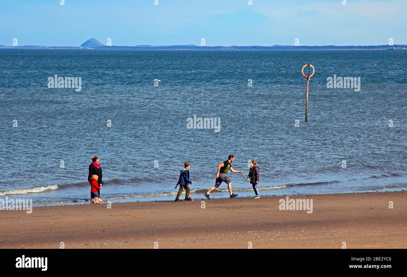 Portobello Promenade, Edinburgh, Scotland, UK. 11th April 2020. Temperature of 16 degrees with full sun in the afternoon after a cloudy start. On Easter Saturday a very quiet beach on the third weekend of Coronavirus Lockdown. A family have fun at the seashore. Stock Photo