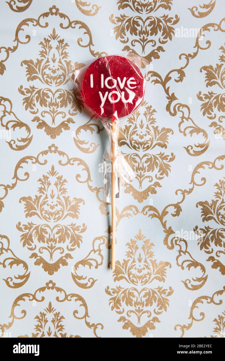 a sweet wrapped 'I Love you' red lollypop against a gold and light blue decorated background, flat lay, top view, minimalistic Stock Photo